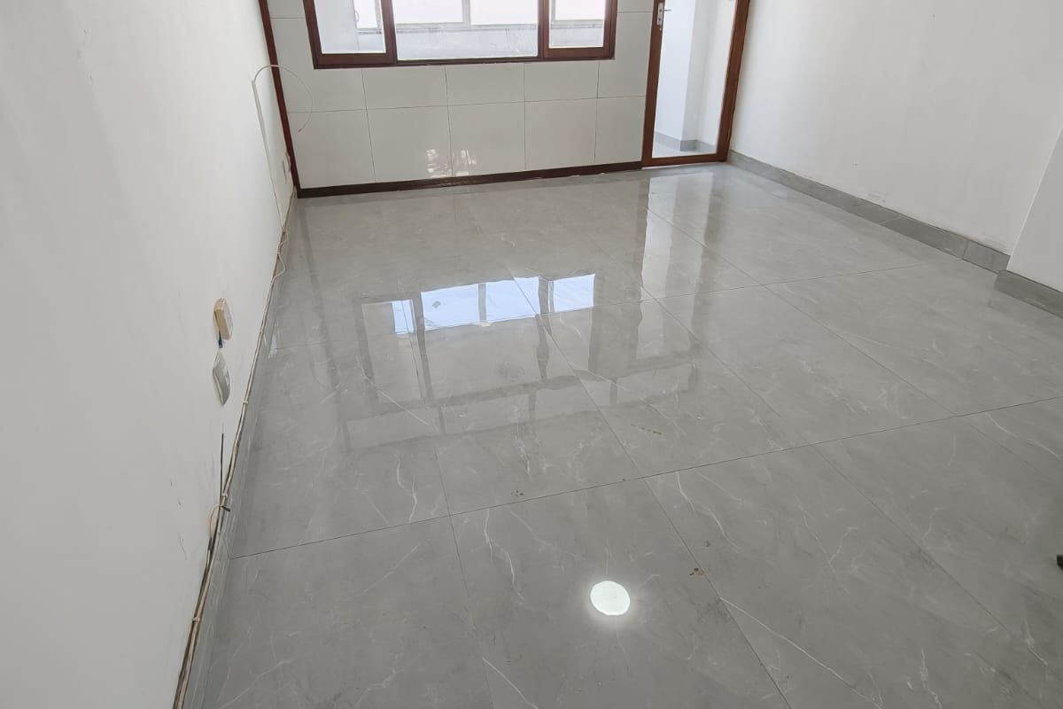 An empty, bright living area featuring modern grey tiled flooring, large windows with aluminium frames, and a glass door leading to an outdoor space.