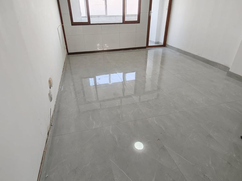An empty, bright living area featuring modern grey tiled flooring, large windows with aluminium frames, and a glass door leading to an outdoor space.