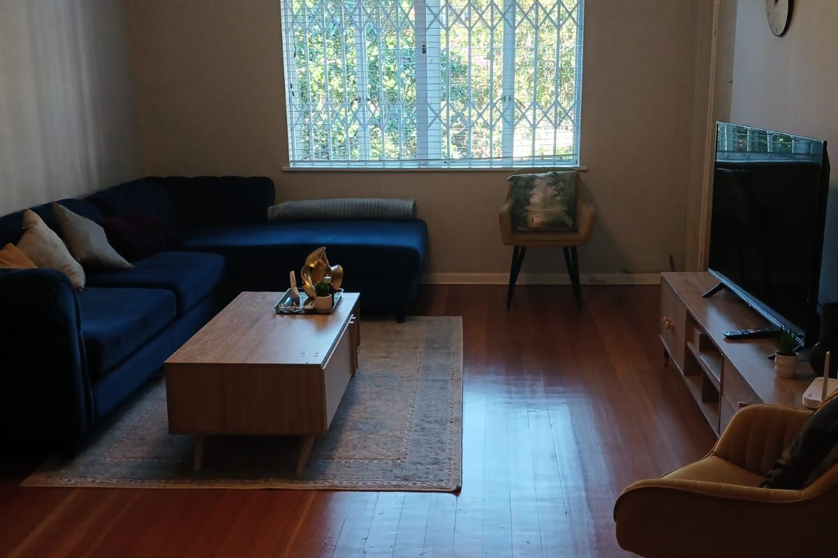 A well-maintained living room featuring polished hardwood flooring, crown molding, a ceiling fan, and a large window fitted with a security gate.