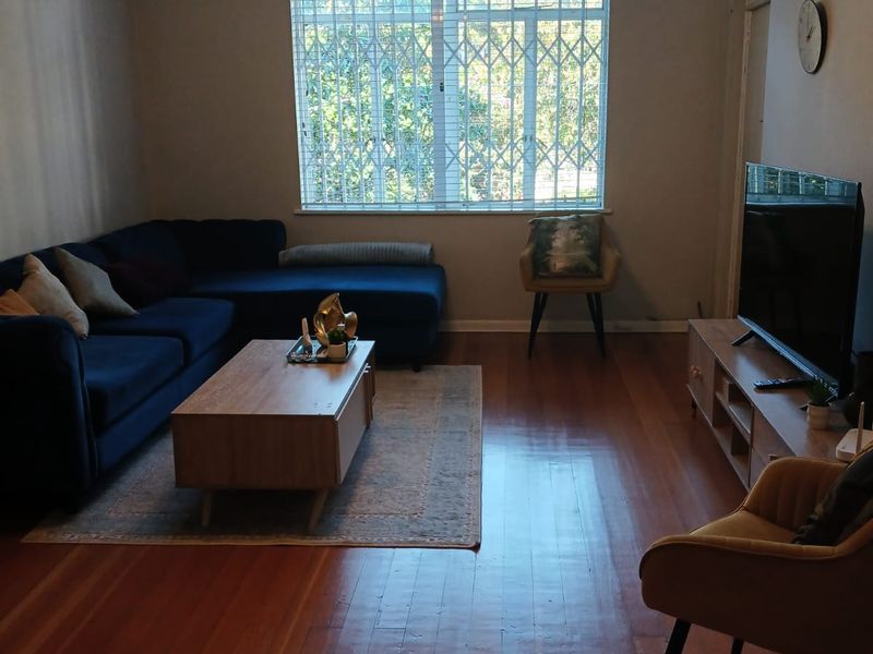 A well-maintained living room featuring polished hardwood flooring, crown molding, a ceiling fan, and a large window fitted with a security gate.