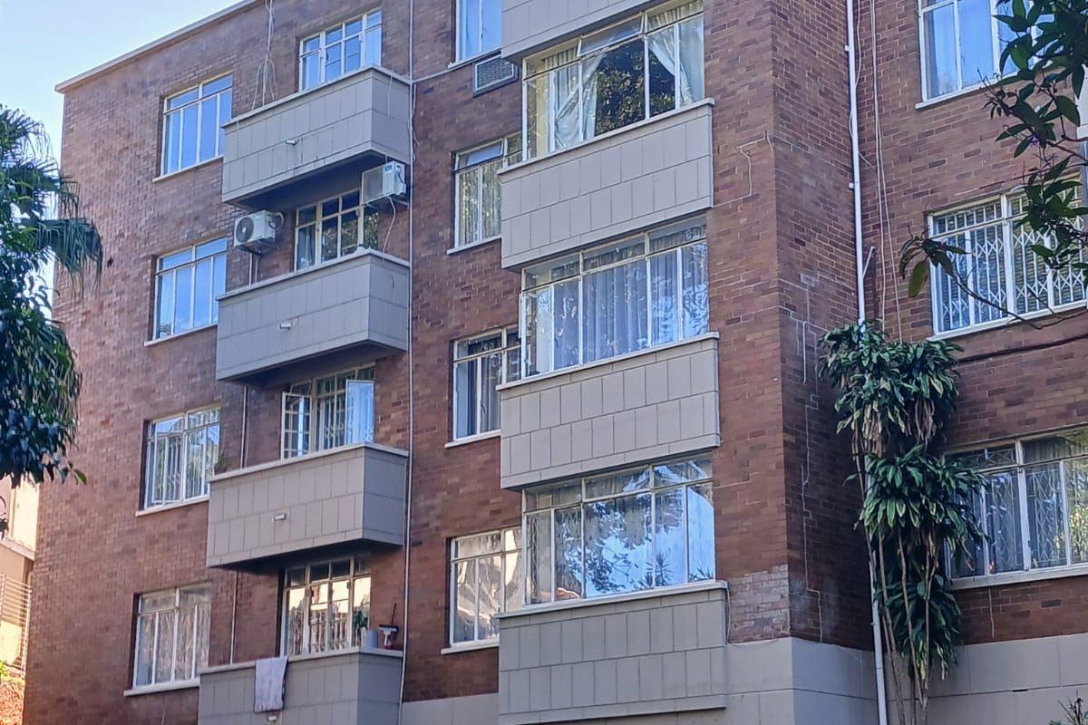 Exterior view of a multi-story brick apartment building featuring private balconies, casement windows, and established lawn area.