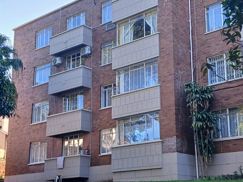 Exterior view of a multi-story brick apartment building featuring private balconies, casement windows, and established lawn area.