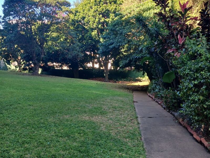 A well-maintained garden area featuring a lush green lawn, mature trees, and a concrete pathway leading through the landscaped grounds.