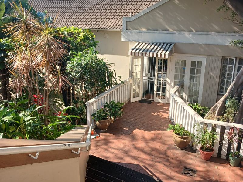 A rear exterior view of the apartment featuring a brick-paved walkway, white railings, and a shaded entrance with a striped awning.