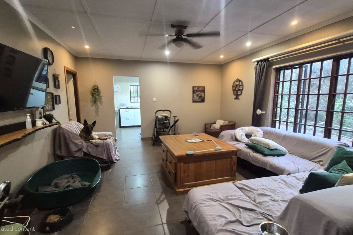 A spacious living room featuring tiled flooring, a ceiling fan, and large windows, with an open flow leading into the adjacent kitchen area.