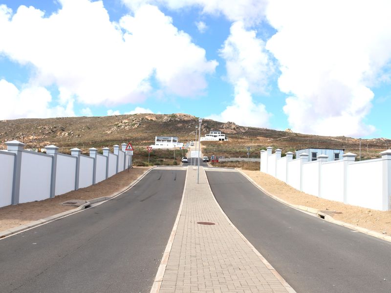 A clear street view of a residential development featuring paved roads, a central brick median, and well-maintained perimeter boundary walls leading towards the hillside.