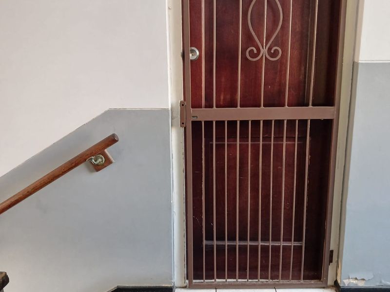 Apartment entrance featuring a wooden door with a security gate, adjacent window, and wall-mounted handrail in a stairwell area.