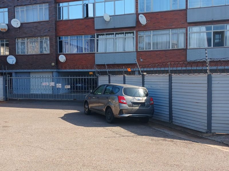 Front exterior view of a multi-story brick apartment building featuring metal security fencing, razor wire, and multiple satellite dishes.