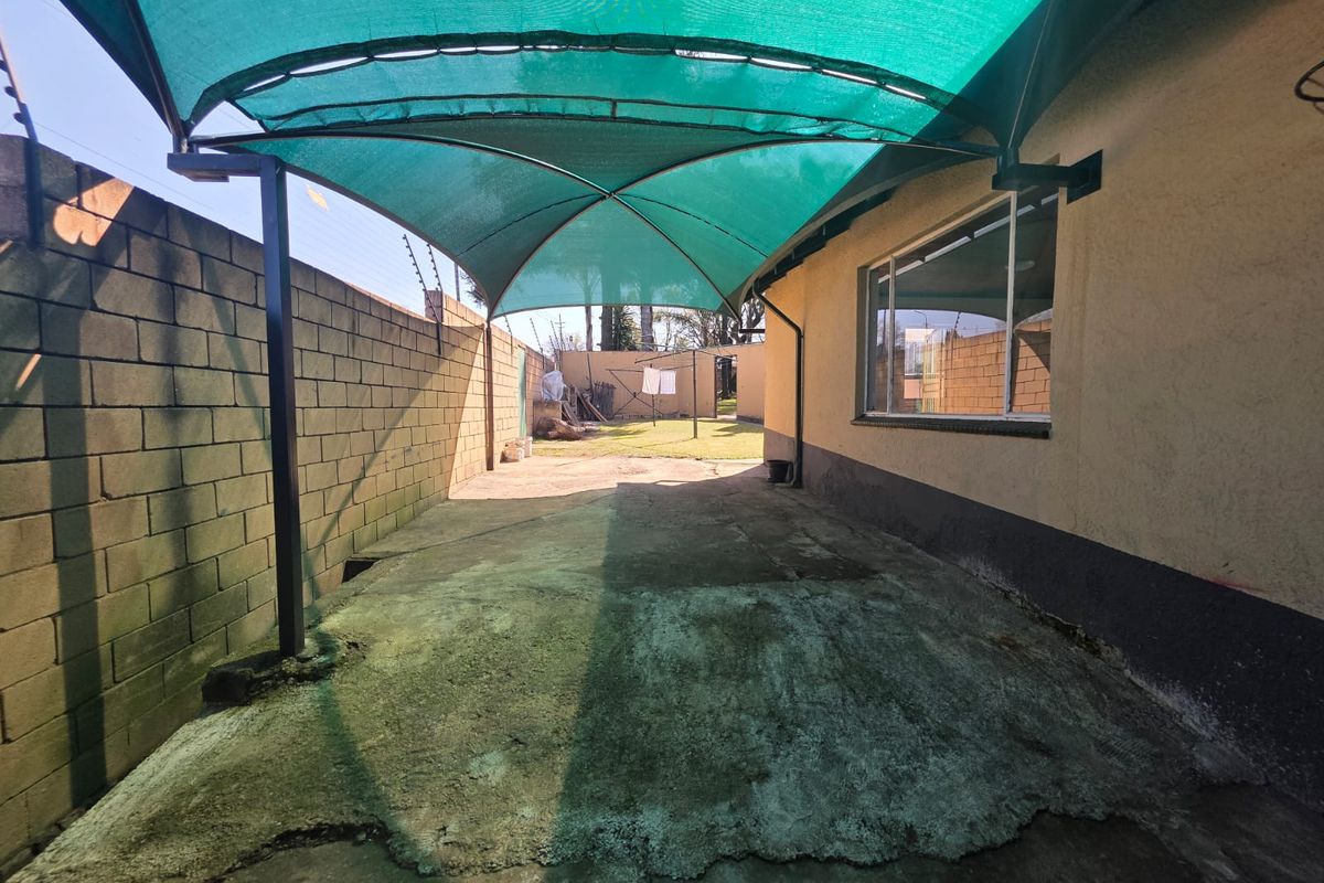 Rear exterior area featuring a green shade sail covering a concrete paved space, enclosed by a boundary wall with electric fencing.