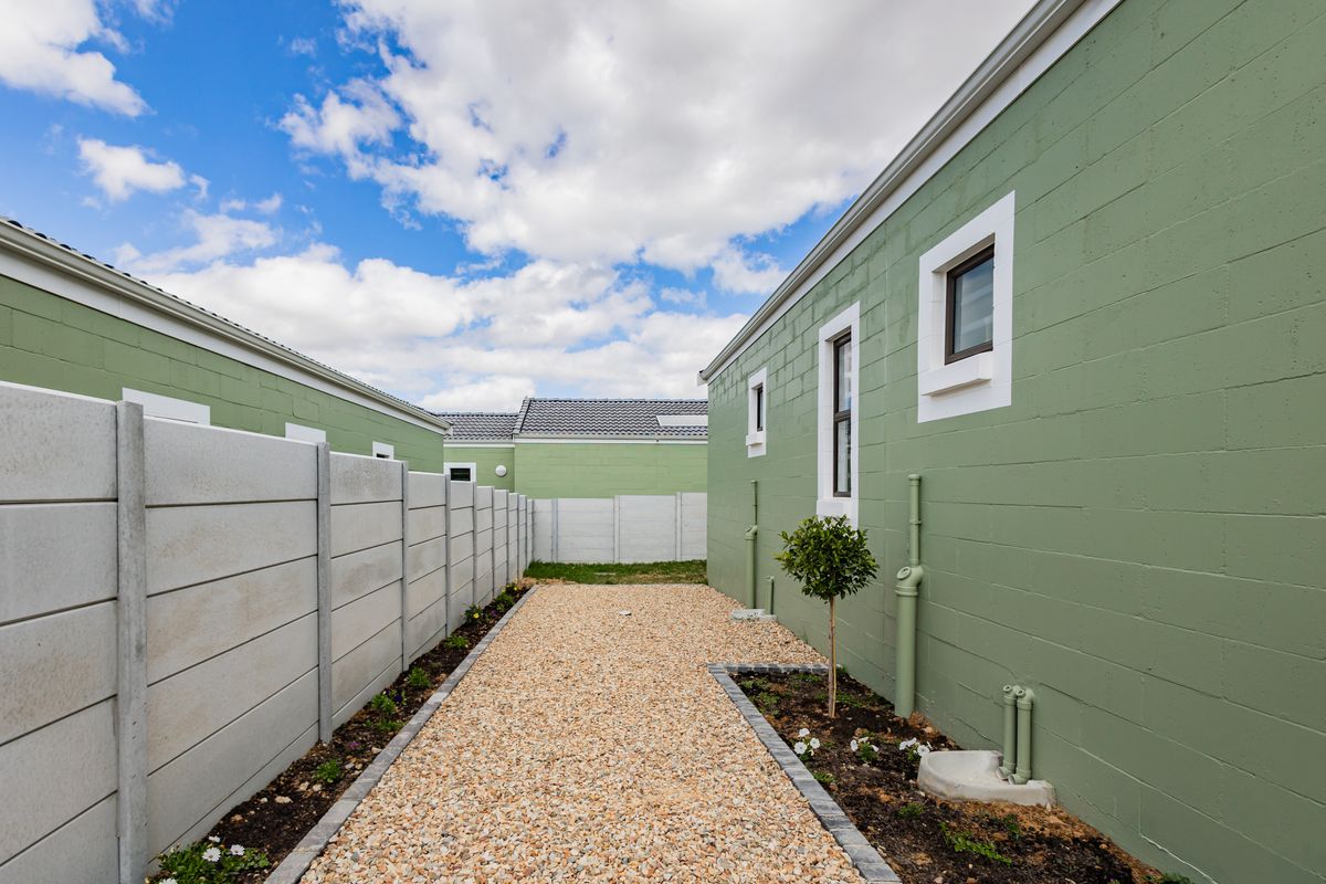 A side exterior view showing a gravel pathway between a green painted concrete block house wall and a precast concrete boundary fence.
