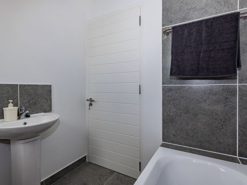 A clean, modern bathroom featuring a pedestal washbasin, a bathtub with grey tiled wall cladding, and neutral floor tiles.