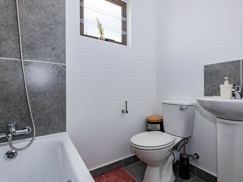 A clean and modern bathroom featuring a bathtub with shower mixer, pedestal basin, and toilet, complemented by grey tiled splashbacks and flooring.