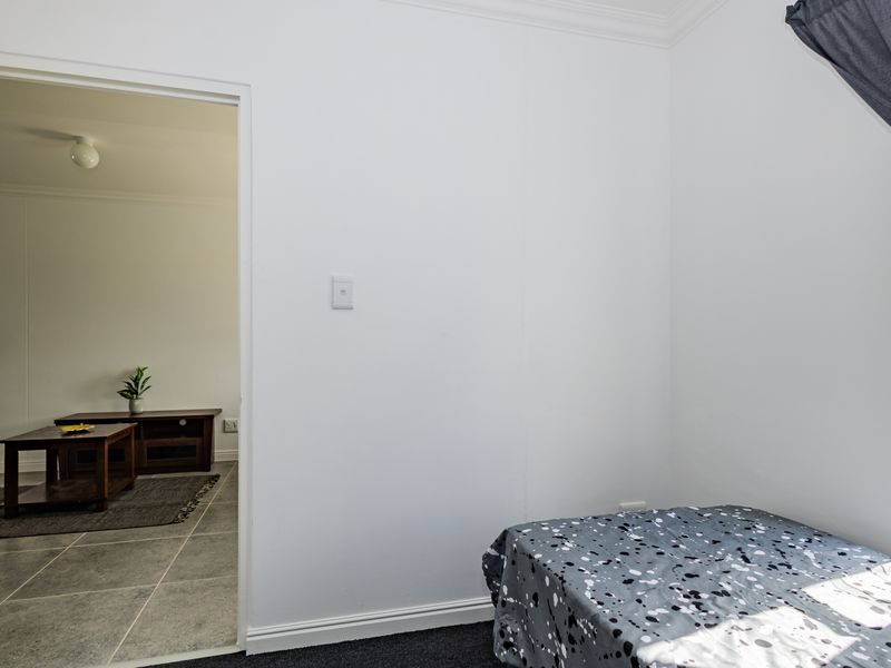 A bedroom featuring neutral white walls, dark carpeting, and a window, with an open doorway leading into an adjacent living area.