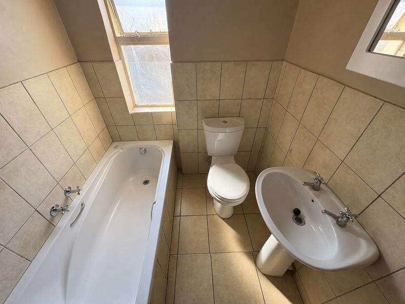 A functional bathroom featuring a standard bathtub, pedestal basin, and toilet with neutral wall and floor tiling.