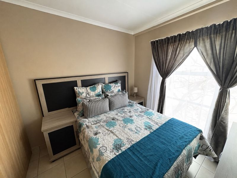 A compact bedroom featuring a built-in headboard with integrated bedside tables, tiled flooring, and a large window with curtain rails.