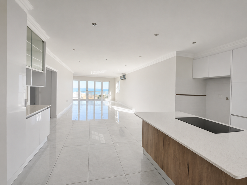 Spacious open-plan living and kitchen area featuring modern cabinetry, a central island with a built-in hob, tiled floors, and large sliding doors to the balcony.