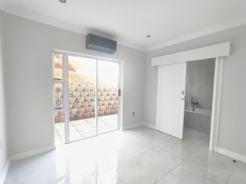 A bright, modern bedroom featuring tiled floors, an air conditioning unit, and a sliding door leading to an outdoor area, with an en-suite bathroom.