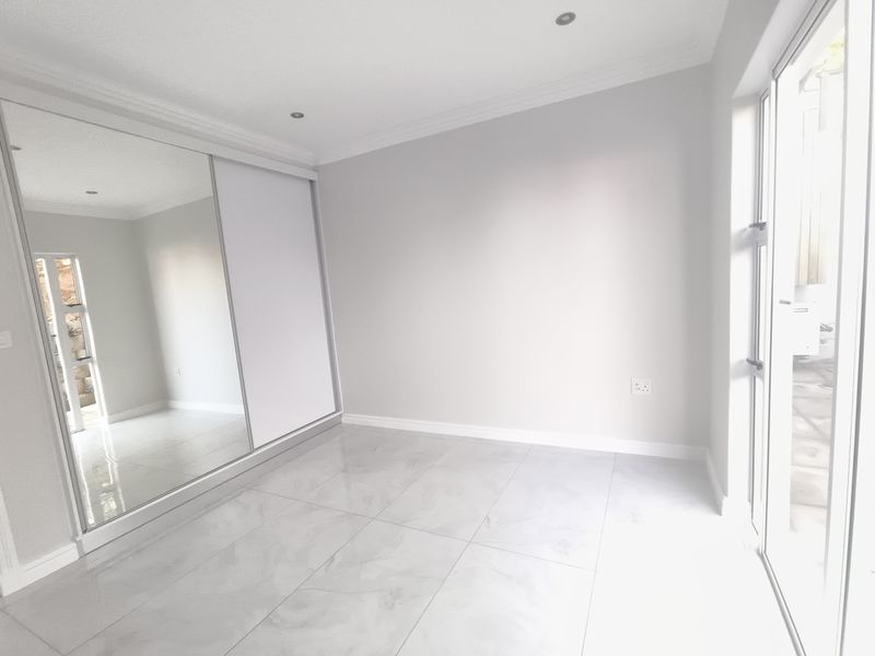A bright, empty bedroom featuring modern tiled flooring, recessed downlights, and a large mirrored sliding wardrobe with direct access to an outdoor area.