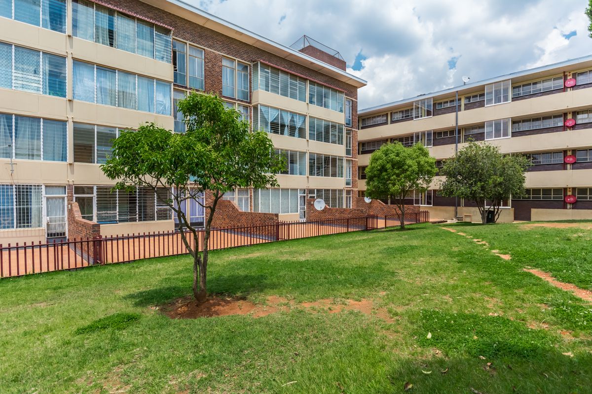 Exterior view of a multi-story apartment complex featuring brick facades, enclosed balconies, and a shared communal garden area with established trees.