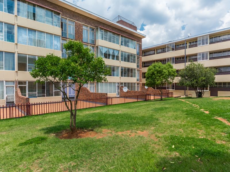 Exterior view of a multi-story apartment complex featuring brick facades, enclosed balconies, and a shared communal garden area with established trees.