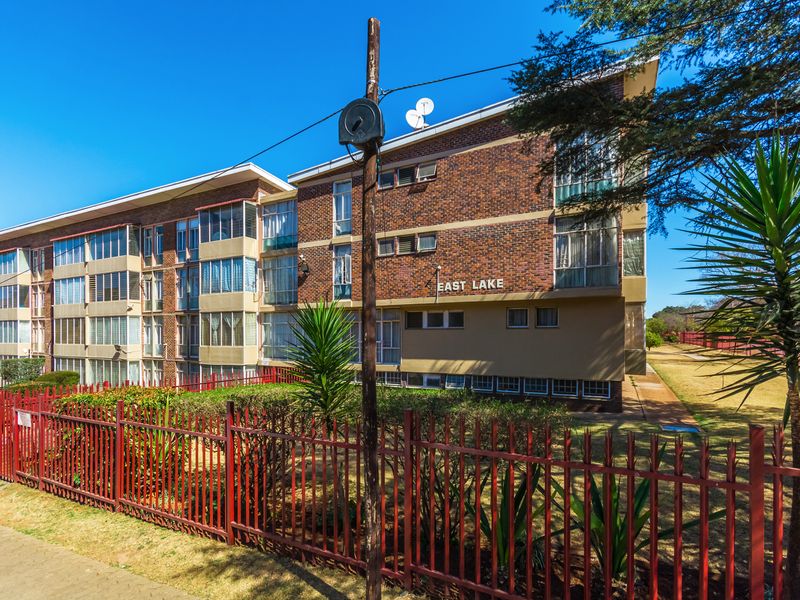 Exterior view of the East Lake apartment building featuring a face brick facade, metal security fencing, and established garden surroundings.