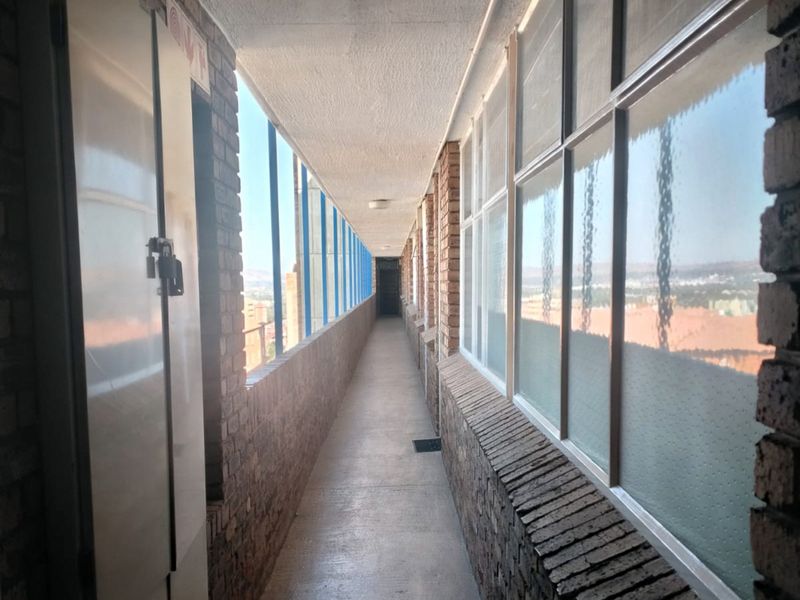 An open-air apartment building hallway featuring exposed brick walls, concrete flooring, and metal-framed windows overlooking the surrounding area.