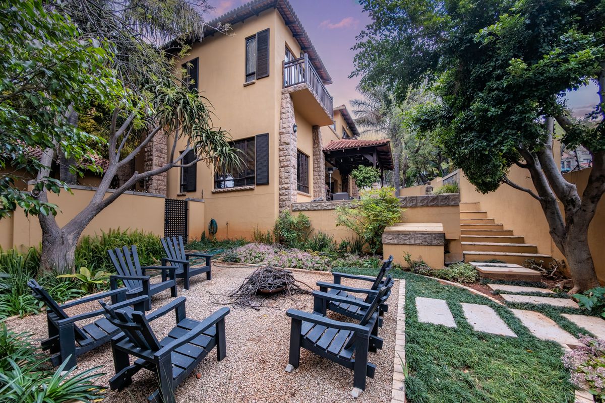 A beautifully landscaped rear garden featuring a gravel fire pit area, stone retaining walls, stairs, and a multi-story home with stone accents and a balcony.