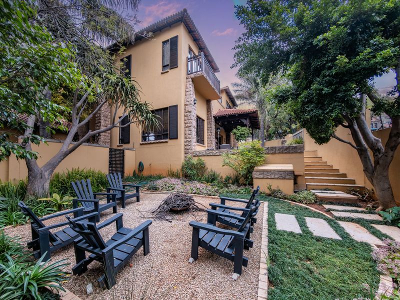 A beautifully landscaped rear garden featuring a gravel fire pit area, stone retaining walls, stairs, and a multi-story home with stone accents and a balcony.