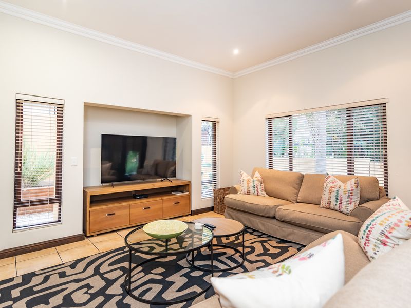 A bright and well-maintained living room featuring tiled flooring, a recessed media wall, and large windows with venetian blinds.