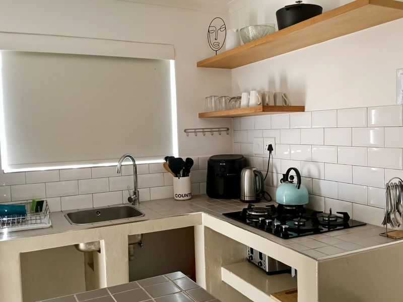 A compact kitchen featuring a tiled countertop, subway tile backsplash, built-in gas cooktop, stainless steel sink, and open timber shelving for storage.