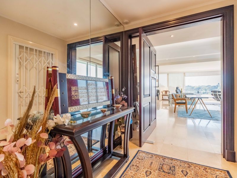 A bright, well-maintained entrance hallway featuring tiled flooring, recessed lighting, and a mirrored wall panel, leading into an open-plan living area.