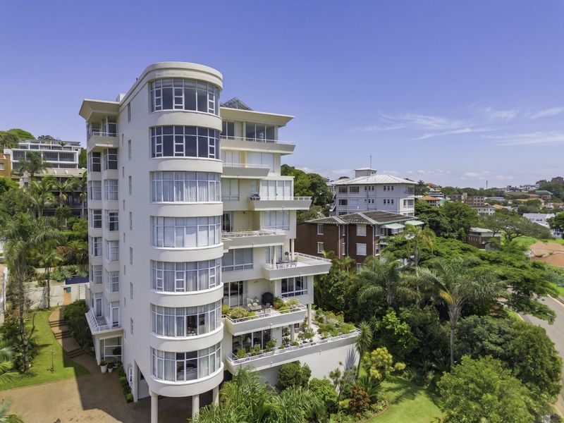 An elevated aerial view of a modern multi-story penthouse building featuring curved balconies, large glass windows, and surrounding landscaped gardens.