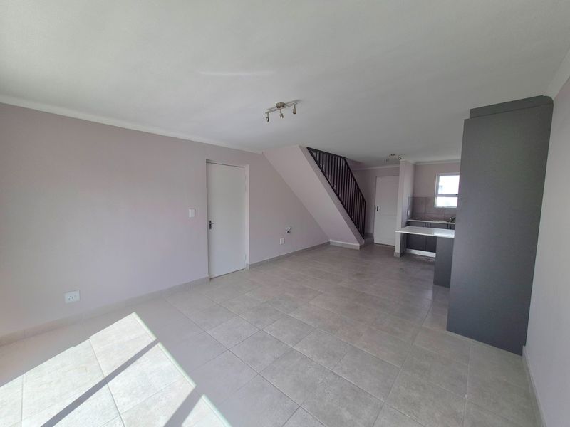Spacious open-plan living area featuring neutral tiled flooring, a modern staircase, and a compact kitchen with built-in cabinetry and countertops.