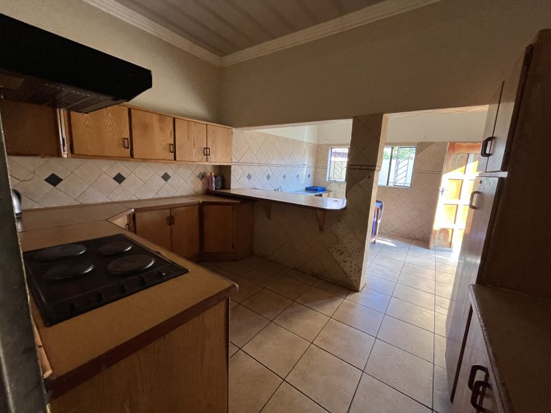 A functional kitchen featuring wooden cabinetry, an electric hob with extractor, tiled backsplash, and a breakfast bar opening into an adjacent living area.