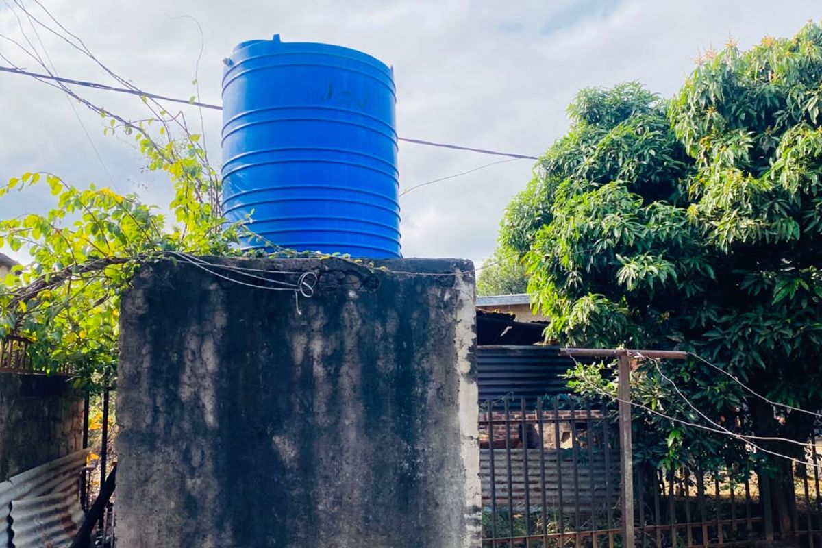 An outdoor area featuring a blue water storage tank mounted on a weathered concrete support structure with surrounding metal fencing and mature trees.