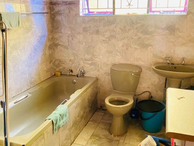 A dated bathroom featuring a bathtub, pedestal basin, and toilet with full-height wall tiling and security bars on the window.