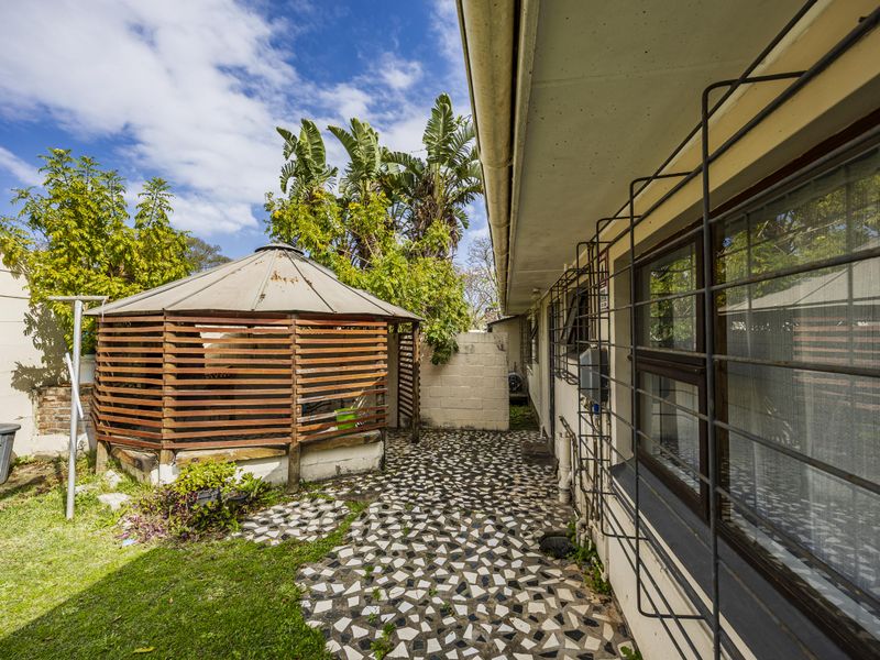 Rear exterior view featuring a wooden gazebo, mosaic paved patio area, and security-barred windows along the side of the house.