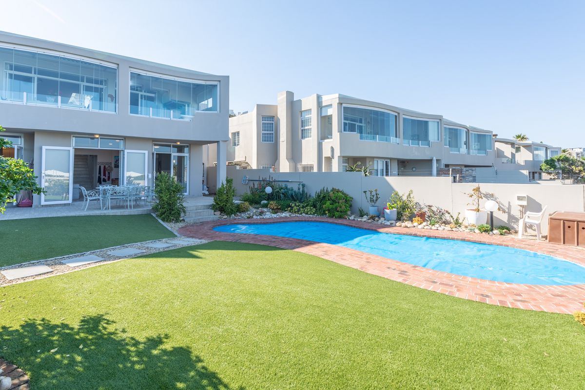 Rear exterior view featuring a swimming pool with brick coping, artificial lawn, paved patio area, and multi-level building with glass-enclosed balconies.