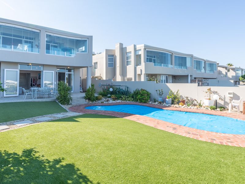 Rear exterior view featuring a swimming pool with brick coping, artificial lawn, paved patio area, and multi-level building with glass-enclosed balconies.
