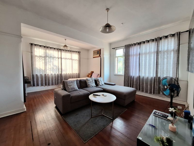Spacious living room featuring original hardwood flooring, large windows with security bars, and a built-in air conditioning unit.