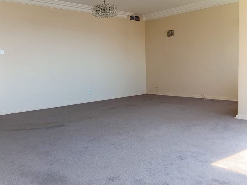 A spacious, empty living room featuring neutral walls, carpeted flooring, decorative crown molding, and a classic ceiling rose with a central light fixture.