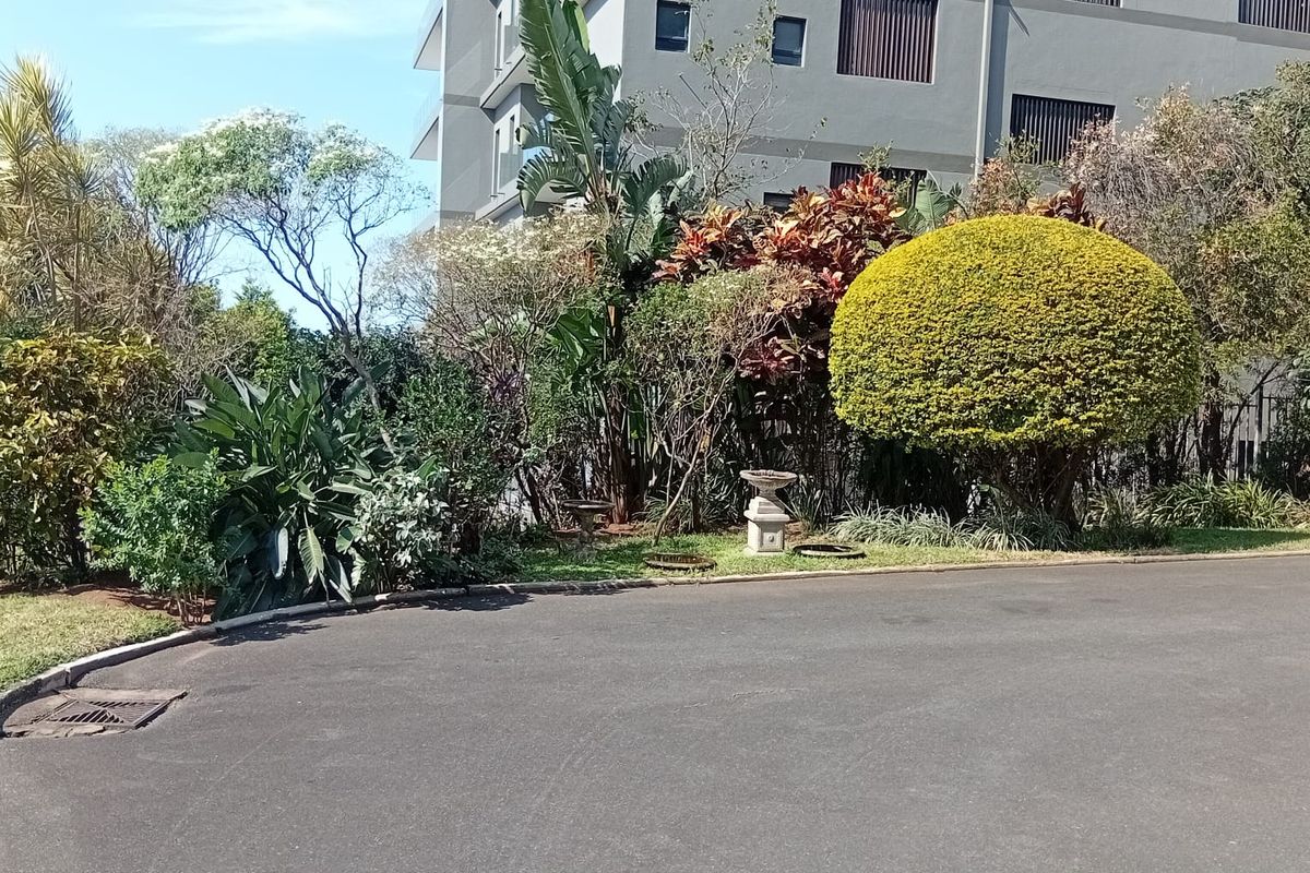 A view of the apartment building exterior featuring a well-maintained landscaped garden and an asphalt driveway with a drainage grate.