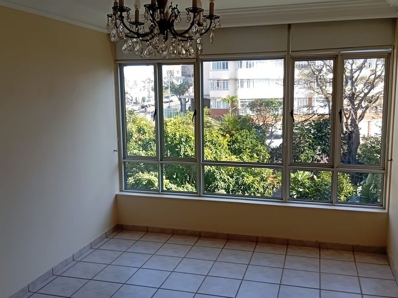 A bright, empty living area featuring tiled flooring, a decorative crystal chandelier, and large windows offering a pleasant leafy outlook.