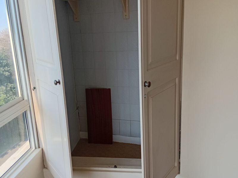 A bedroom featuring a built-in cupboard with tiled interior shelving and a large window, showing signs of wear around the door frame.