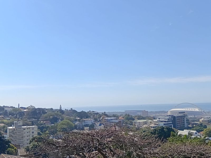 Elevated panoramic view overlooking the surrounding neighborhood, coastal landscape, and distant stadium structure under a clear blue sky.
