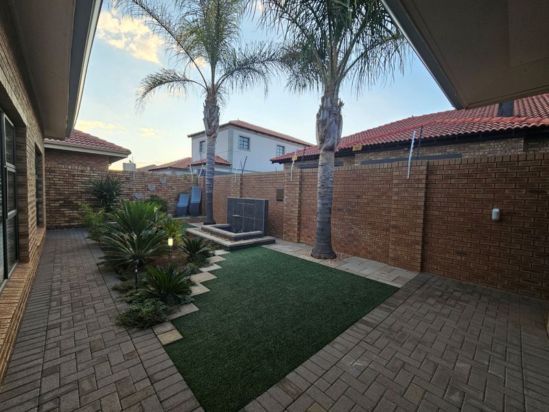 A well-maintained rear garden featuring a paved patio area, artificial lawn, a built-in water feature, and secure brick boundary walls with electric fencing.