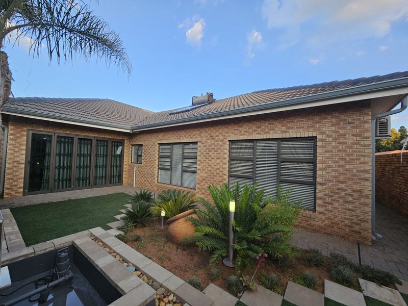 Rear exterior view featuring a brick facade, tiled roof with solar geyser, aluminium windows with security gates, and a landscaped garden with a water feature.