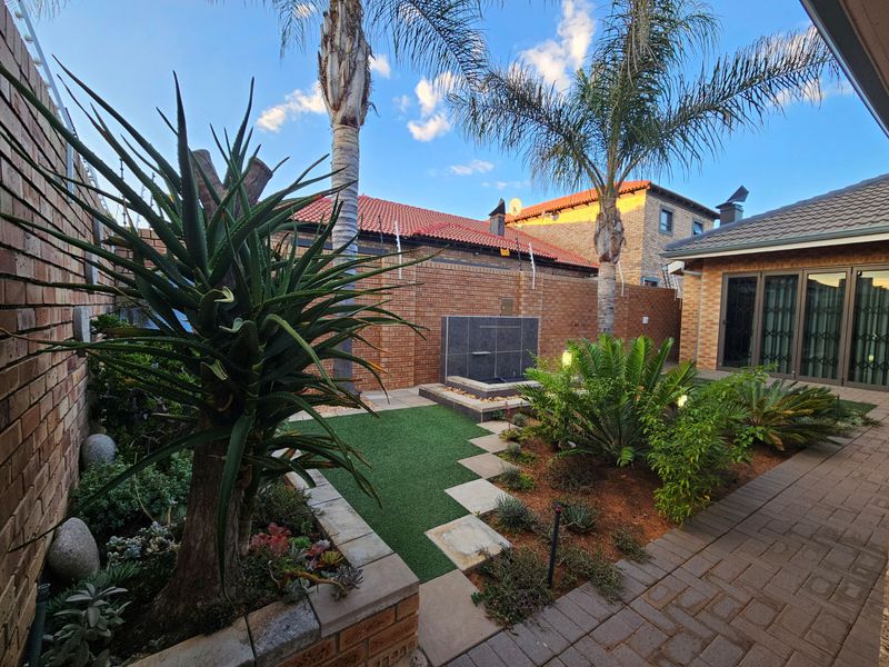 A well-maintained rear courtyard featuring a paved patio, artificial lawn, landscaped garden beds, and a modern water feature.