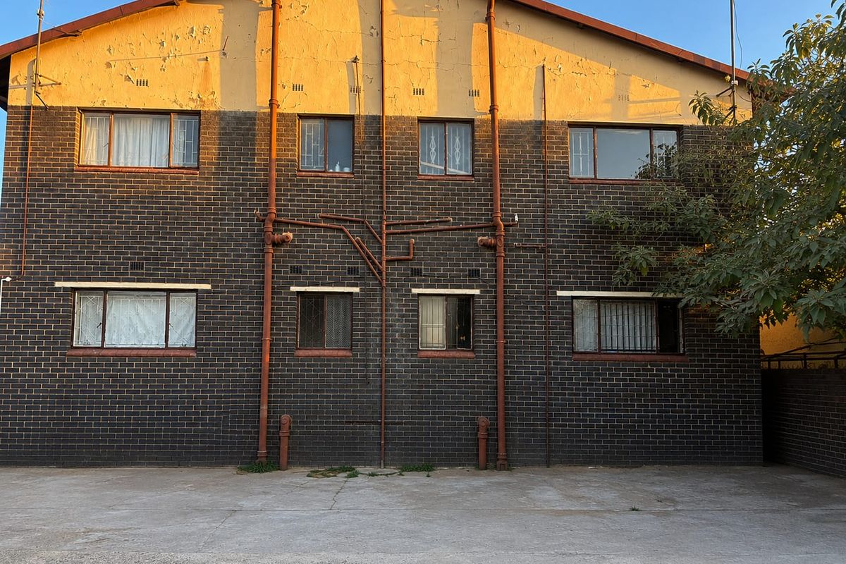 Rear elevation of a multi-story brick apartment building featuring exposed plumbing, security bars on windows, and a concrete ground area.