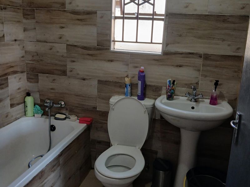 A functional bathroom featuring a bathtub, pedestal basin, and toilet, finished with wood-look wall tiles and security bars on the window.